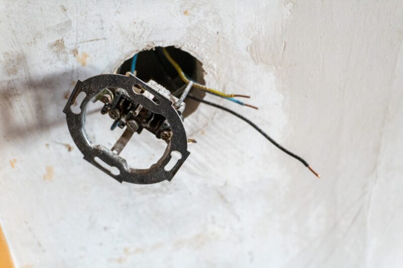 An exposed electrical wall socket emerges from a rough white plaster wall, with loose wires extending outward, capturing a raw moment of home renovation and unfinished construction. The minimal, industrial scene reflects maintenance, repair work, and the hidden infrastructure behind modern interiors.